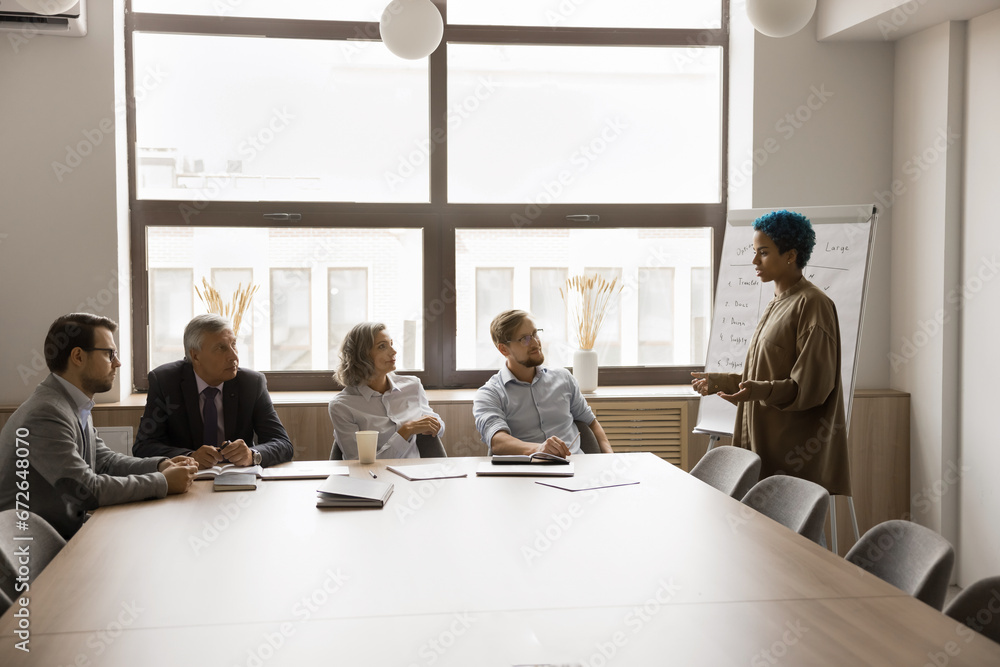 Fototapeta premium Young African American creative manager girl talking to diverse coworkers, presenting sales report, brainstorming idea, project plan, marketing strategy, speaking at whiteboard on meeting