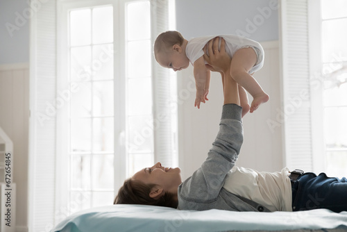 Side view loving mother lying on comfortable bed in cozy, light bedroom play with infant, lifts un outstretched arms her adorable awakened newborn smile, enjoy sweet moments together at home. Infancy
