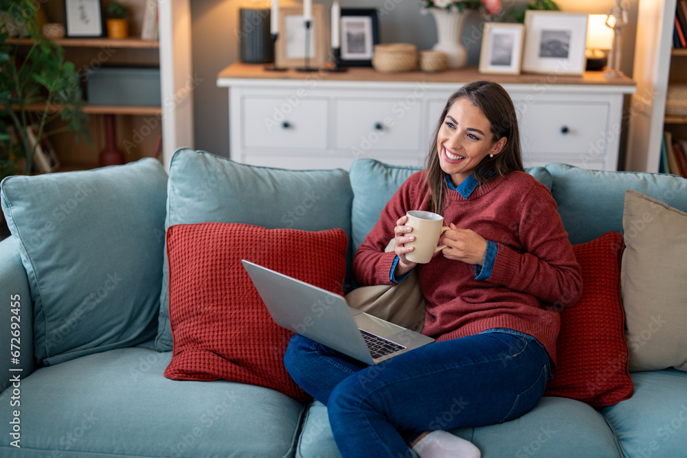 Obraz premium Smiling woman freelancer sitting on sofa in home office with coffee cup and laptop.