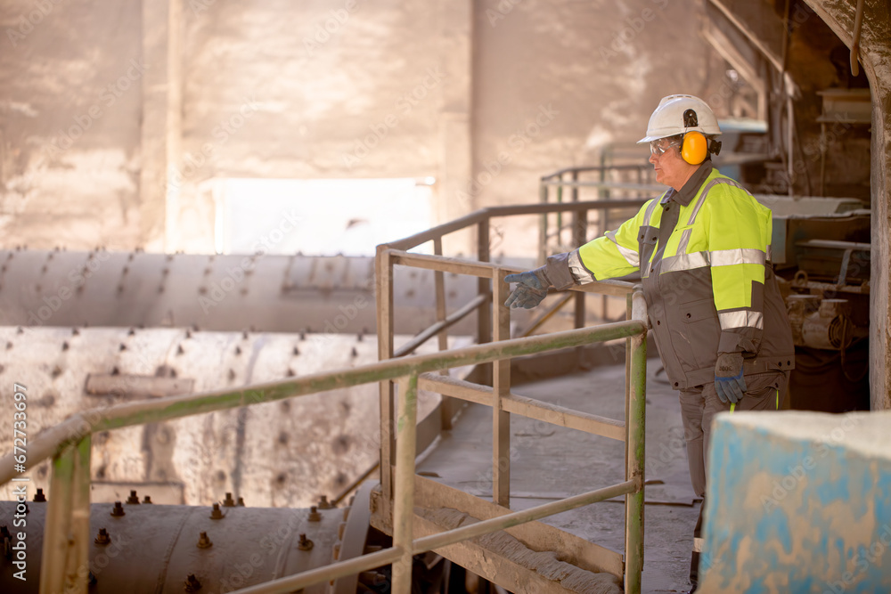 A worker in a helmet and equipment stands against the backdrop of a cement plant workshop. Work technology, cement production. People working in heavy industry. A worker in a helmet and equipment stands against the backdrop of a cement plant workshop. Work technology, cement production. People working in heavy industry.
