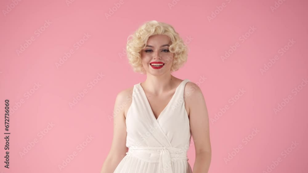 Portrait young cheerful woman in wig, white dress and with red lipstick on lips in studio on pink background. Woman portrays actress , copying her movements, fixing her hair. Portrait young cheerful woman in wig, white dress and with red lipstick on lips in studio on pink background. Woman portrays actress , copying her movements, fixing her hair.