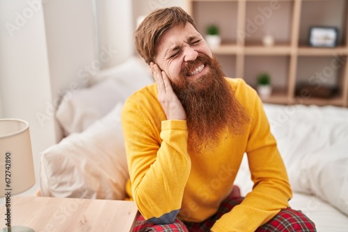 Young redhead man suffering for ear pain sitting on bed at bedroom