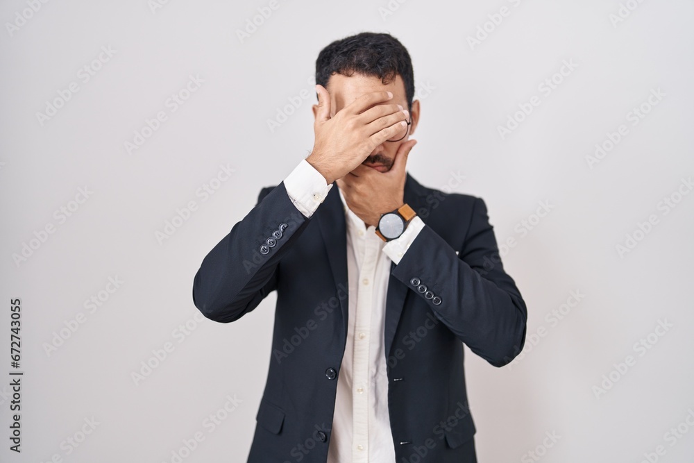 Handsome business hispanic man standing over white background covering eyes and mouth with hands, surprised and shocked. hiding emotion Handsome business hispanic man standing over white background covering eyes and mouth with hands, surprised and shocked. hiding emotion