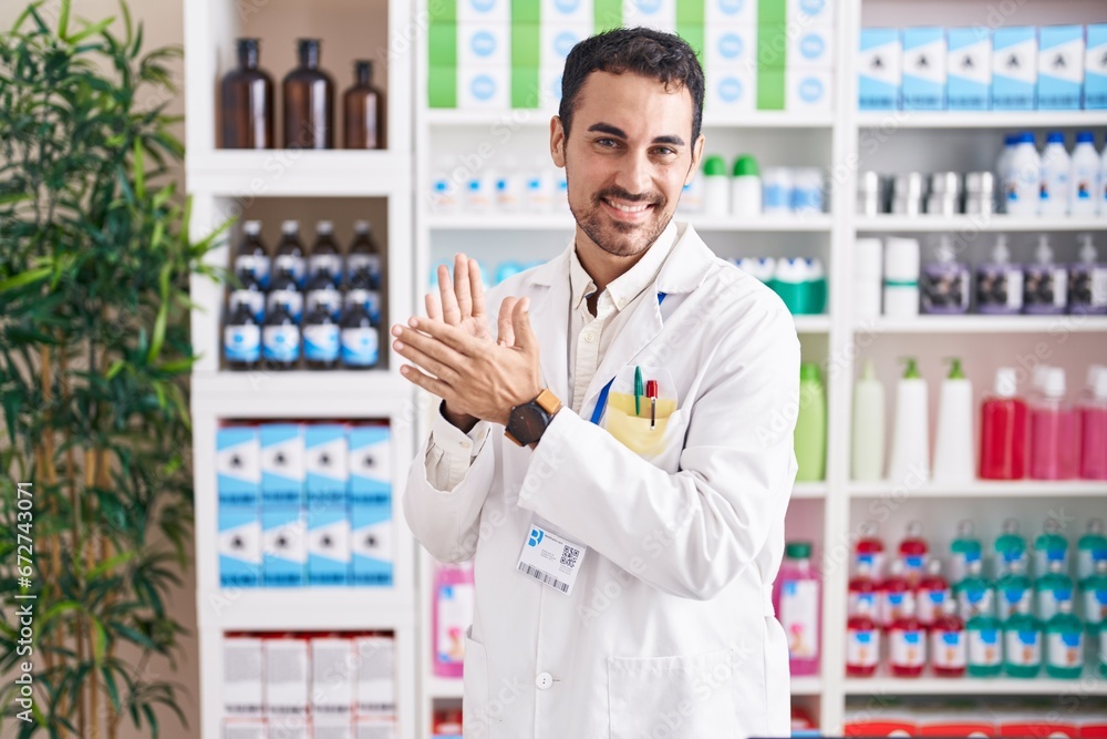 Handsome hispanic man working at pharmacy drugstore clapping and applauding happy and joyful, smiling proud hands together