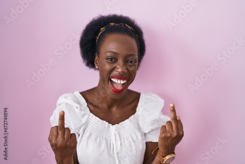 African woman with curly hair standing over pink background showing middle finger doing fuck you bad expression, provocation and rude attitude. screaming excited