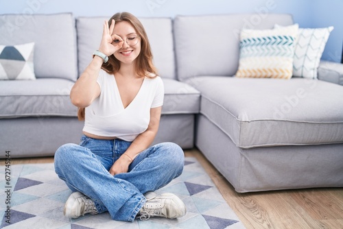 Young caucasian woman sitting on the floor at the living room doing ok gesture with hand smiling, eye looking through fingers with happy face.