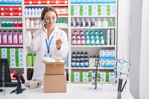 Young blonde woman pharmacist holding pills bottle talking on smartphone at pharmacy