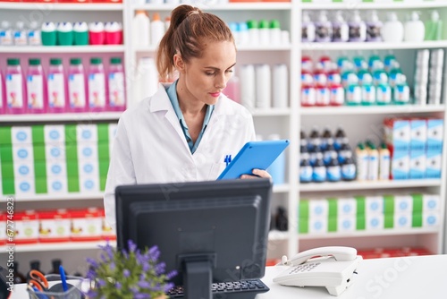 Young beautiful hispanic woman pharmacist using computer and touchpad with serious expression at pharmacy