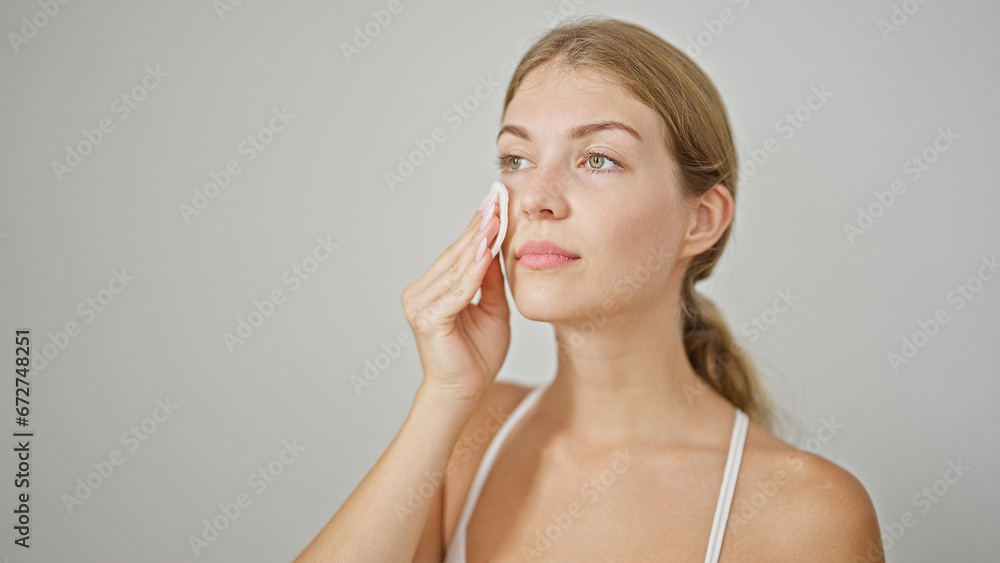 Young blonde woman cleaning face with cotton pad over isolated white background Young blonde woman cleaning face with cotton pad over isolated white background