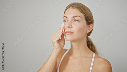 Young blonde woman cleaning face with cotton pad over isolated white background