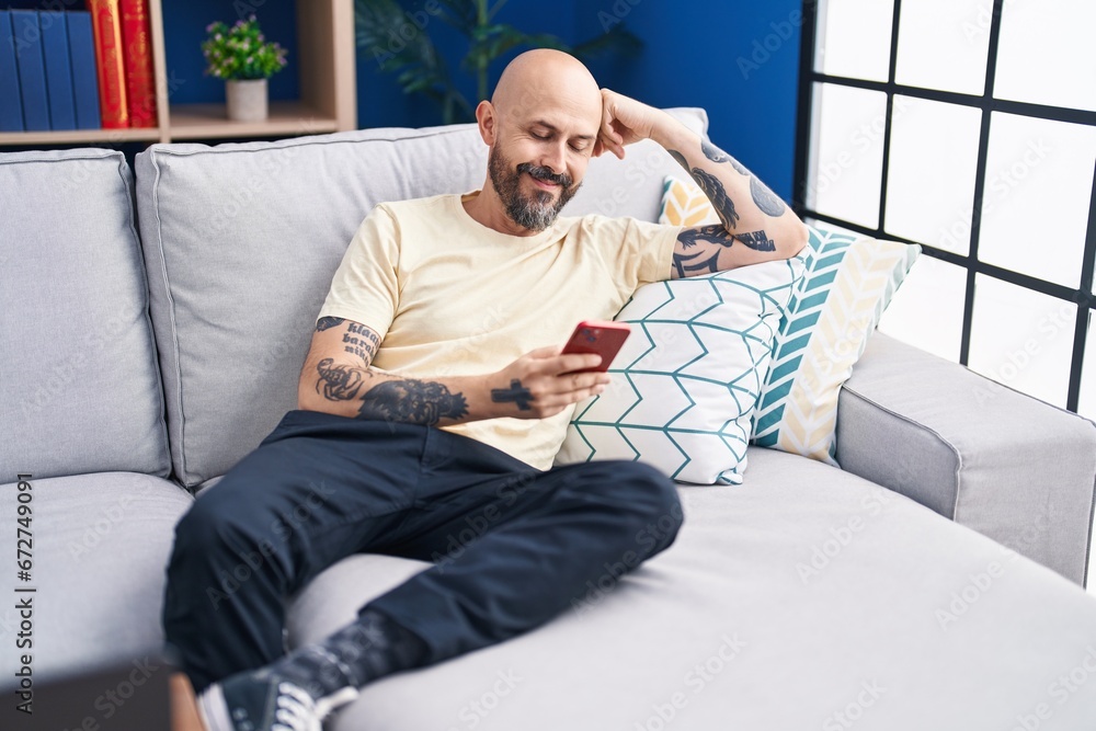 Young bald man using smartphone sitting on sofa at home Young bald man using smartphone sitting on sofa at home