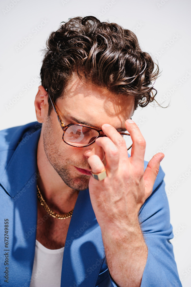 Fashionable brunette man in blue jacket and golden chair touching eyeglasses isolated on grey Fashionable brunette man in blue jacket and golden chair touching eyeglasses isolated on grey