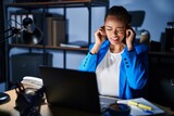 Beautiful african american woman working at the office at night covering ears with fingers with annoyed expression for the noise of loud music. deaf concept.
