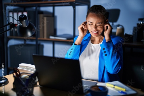 Beautiful african american woman working at the office at night covering ears with fingers with annoyed expression for the noise of loud music. deaf concept. photo