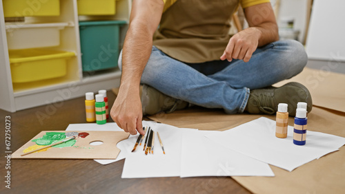 Young latin man artist sitting on floor holding paintbrush at art studio