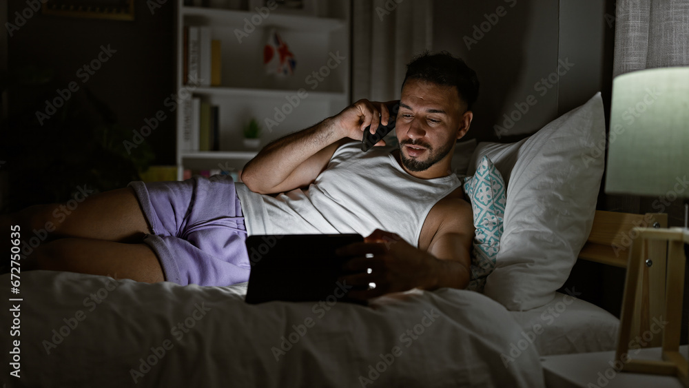 Young arab man talking on smartphone using touchpad lying on bed at bedroom Young arab man talking on smartphone using touchpad lying on bed at bedroom