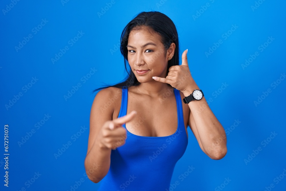 Hispanic woman standing over blue background smiling doing talking on the telephone gesture and pointing to you. call me. Hispanic woman standing over blue background smiling doing talking on the telephone gesture and pointing to you. call me.