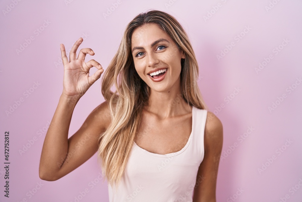 Young blonde woman standing over pink background smiling positive doing ok sign with hand and fingers. successful expression.