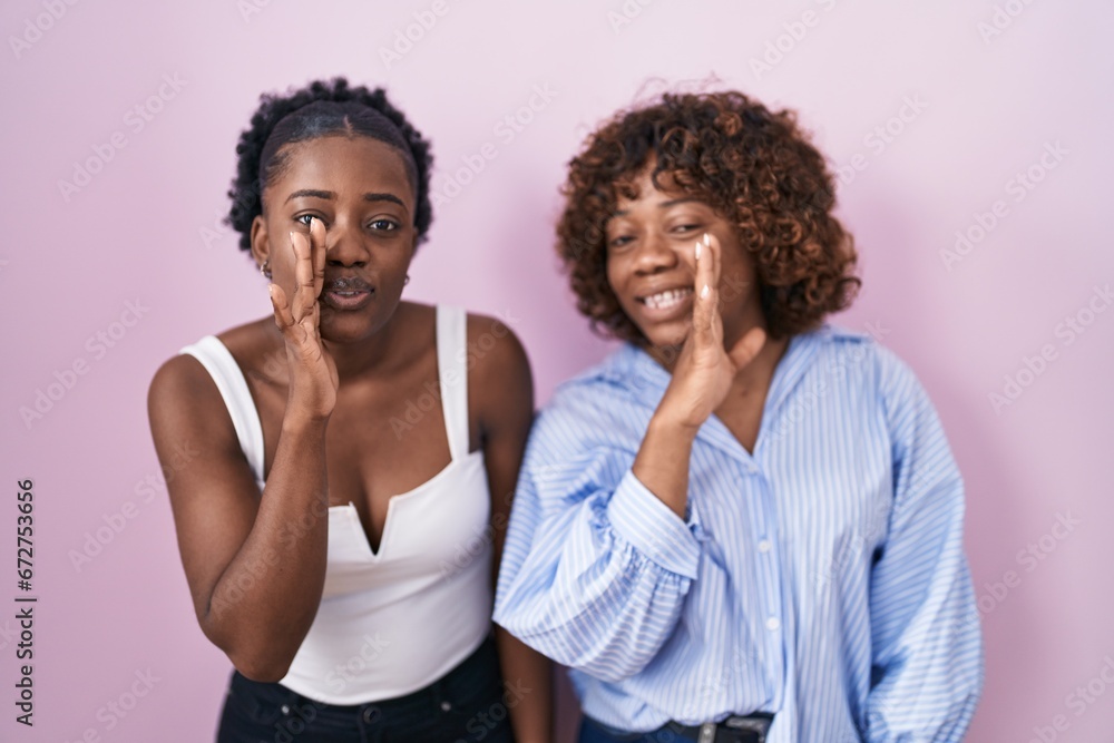 Obraz premium Two african women standing over pink background hand on mouth telling secret rumor, whispering malicious talk conversation