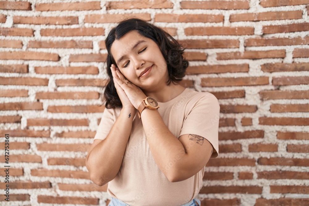 Young hispanic woman standing over bricks wall sleeping tired dreaming and posing with hands together while smiling with closed eyes.