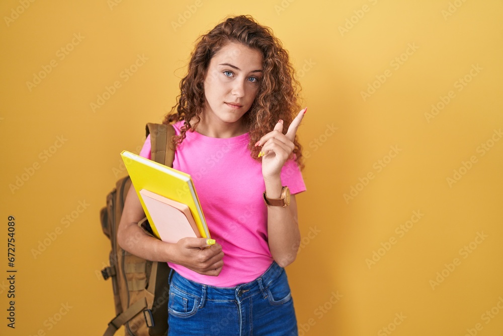 Young caucasian woman wearing student backpack and holding books pointing with hand finger to the side showing advertisement, serious and calm face Young caucasian woman wearing student backpack and holding books pointing with hand finger to the side showing advertisement, serious and calm face