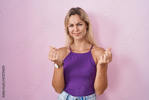 Young blonde woman standing over pink background doing money gesture with hands  asking for salary payment  millionaire business