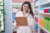 Young caucasian woman pharmacist talking on smartphone reading document at pharmacy