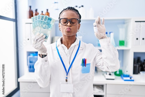 African woman with braids working at scientist laboratory holding money looking at the camera blowing a kiss being lovely and sexy. love expression.