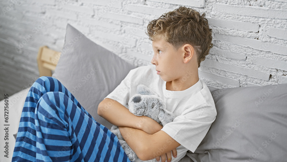 Adorable blond boy in pyjamas, sitting on bed in his bedroom, hugging teddy bear with sad expression, showing signs of unhappiness in the early morning