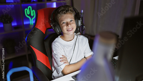 Cute blond boy streamer, engrossed in a digital game, sitting cross-armed and smiling in a dark gaming room lit up by futuristic virtual entertainment technology. © Krakenimages.com