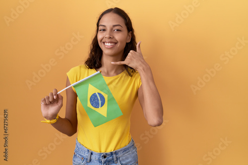 Young hispanic woman holding brazil flag smiling doing phone gesture with hand and fingers like talking on the telephone. communicating concepts.