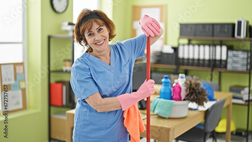 Cuadro en lienzo Middle age woman professional cleaner holding mop stick smiling at office