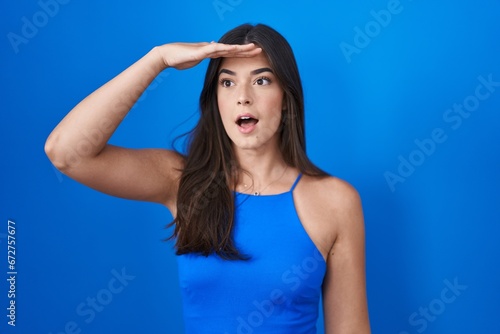 Hispanic woman standing over blue background very happy and smiling looking far away with hand over head. searching concept.