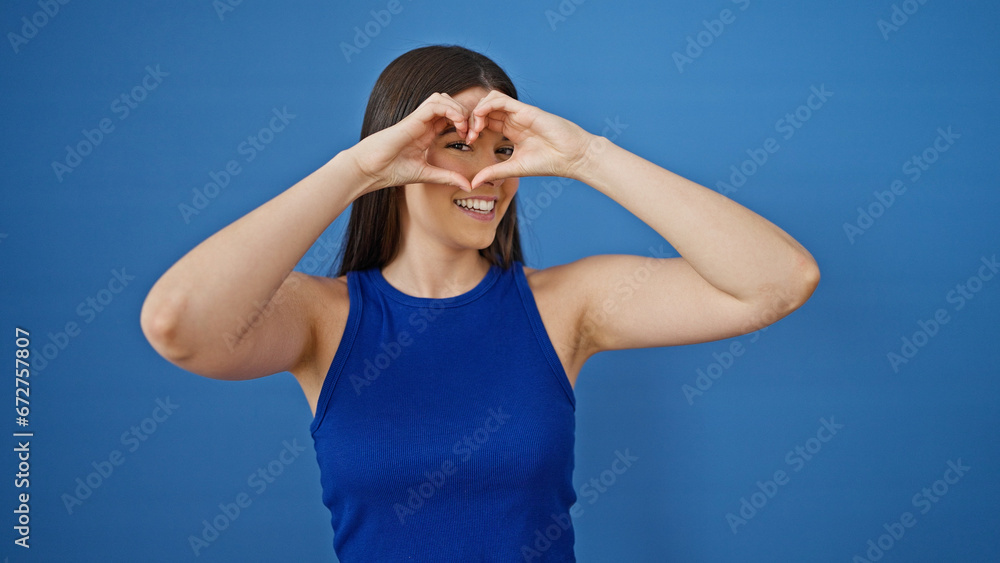 Young beautiful hispanic woman smiling confident doing heart gesture with hands over isolated blue background
