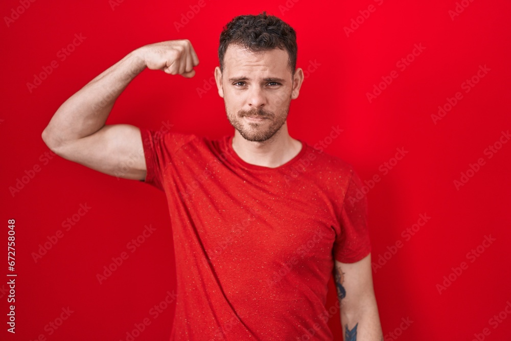 Young hispanic man standing over red background strong person showing arm muscle, confident and proud of power Young hispanic man standing over red background strong person showing arm muscle, confident and proud of power