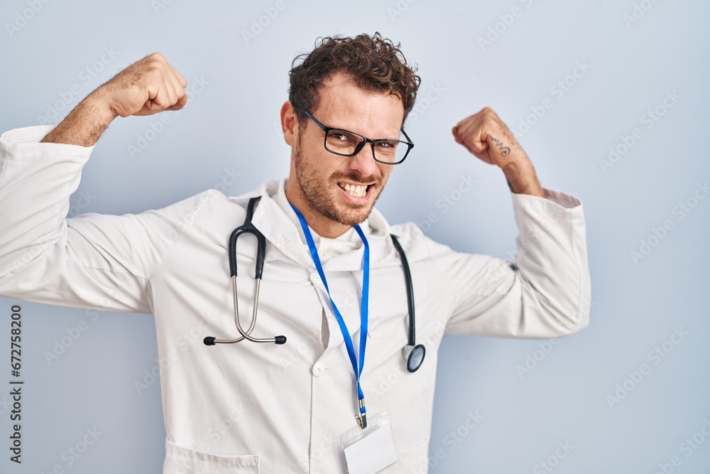 Young hispanic man wearing doctor uniform and stethoscope showing arms muscles smiling proud. fitness concept. Young hispanic man wearing doctor uniform and stethoscope showing arms muscles smiling proud. fitness concept.
