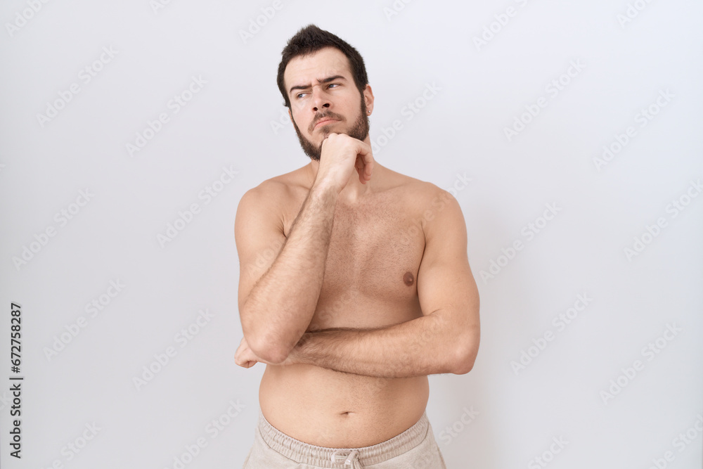 Fototapeta premium Young hispanic man standing shirtless over white background with hand on chin thinking about question, pensive expression. smiling with thoughtful face. doubt concept.