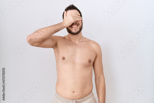 Young hispanic man standing shirtless over white background smiling and laughing with hand on face covering eyes for surprise. blind concept.