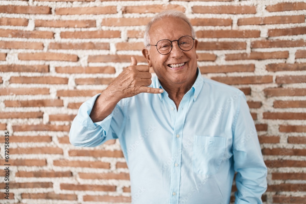 Senior man with grey hair standing over bricks wall smiling doing phone gesture with hand and fingers like talking on the telephone. communicating concepts. Senior man with grey hair standing over bricks wall smiling doing phone gesture with hand and fingers like talking on the telephone. communicating concepts.