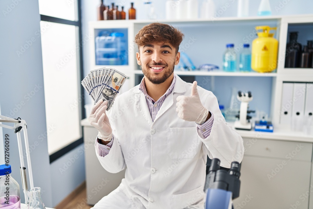 Arab man with beard working at scientist laboratory holding money smiling happy and positive, thumb up doing excellent and approval sign Arab man with beard working at scientist laboratory holding money smiling happy and positive, thumb up doing excellent and approval sign