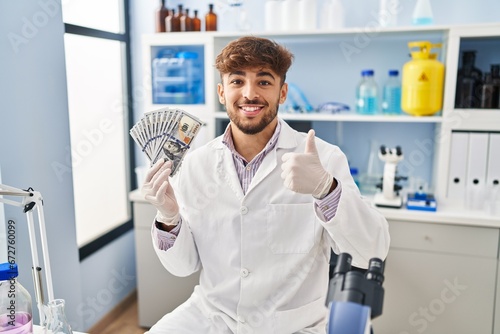 Arab man with beard working at scientist laboratory holding money smiling happy and positive  thumb up doing excellent and approval sign