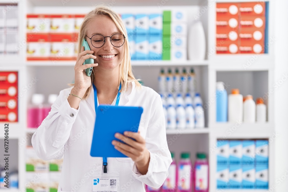 Young blonde woman pharmacist using touchpad talking on smartphone at pharmacy Young blonde woman pharmacist using touchpad talking on smartphone at pharmacy