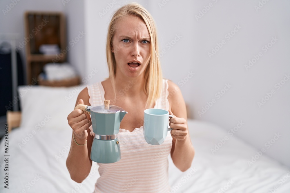 Young caucasian woman drinking coffee on the bed in shock face, looking skeptical and sarcastic, surprised with open mouth Young caucasian woman drinking coffee on the bed in shock face, looking skeptical and sarcastic, surprised with open mouth