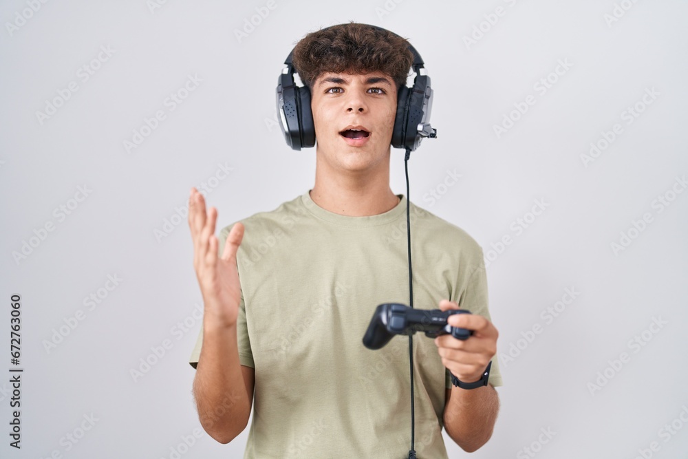 Hispanic teenager playing video game holding controller crazy and mad shouting and yelling with aggressive expression and arms raised. frustration concept. Hispanic teenager playing video game holding controller crazy and mad shouting and yelling with aggressive expression and arms raised. frustration concept.