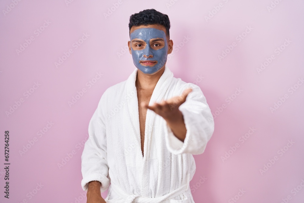 Young hispanic man wearing beauty face mask and bath robe smiling cheerful offering palm hand giving assistance and acceptance.