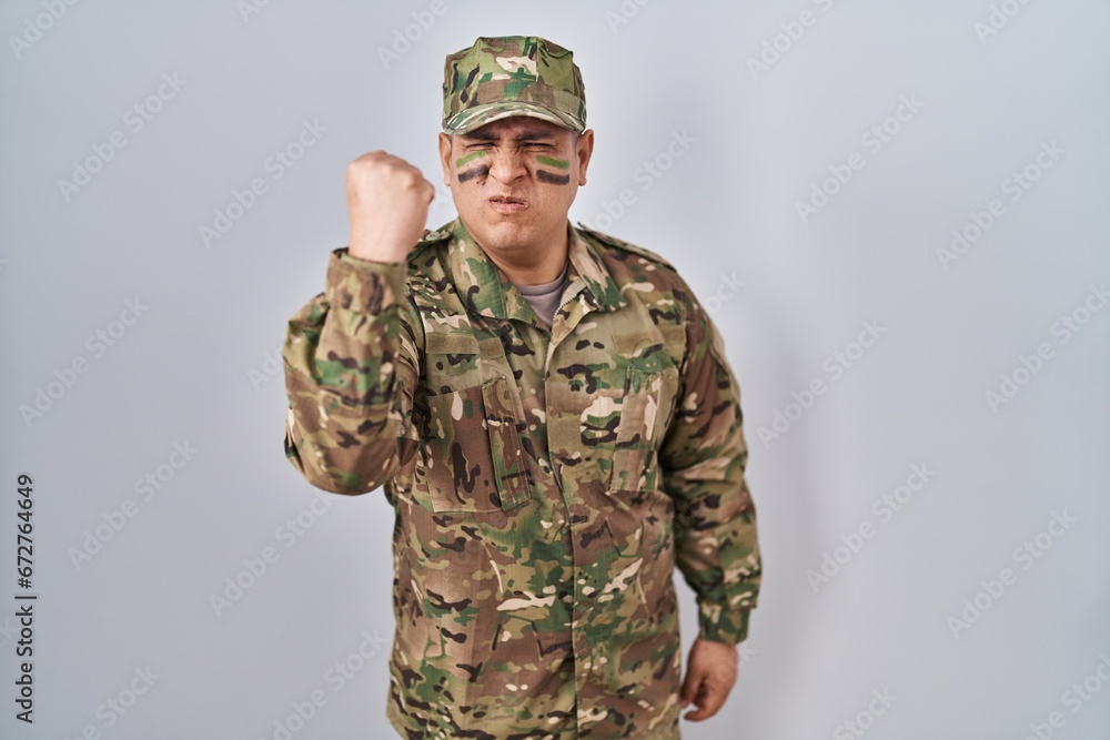Hispanic young man wearing camouflage army uniform angry and mad raising fist frustrated and furious while shouting with anger. rage and aggressive concept. Hispanic young man wearing camouflage army uniform angry and mad raising fist frustrated and furious while shouting with anger. rage and aggressive concept.