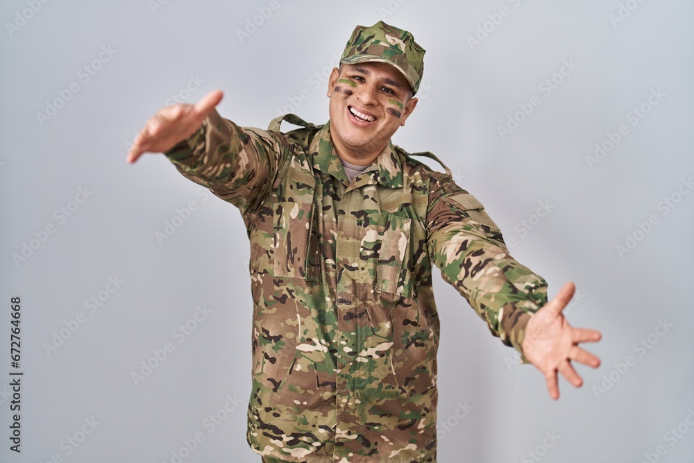 Hispanic young man wearing camouflage army uniform looking at the camera smiling with open arms for hug. cheerful expression embracing happiness. Hispanic young man wearing camouflage army uniform looking at the camera smiling with open arms for hug. cheerful expression embracing happiness.