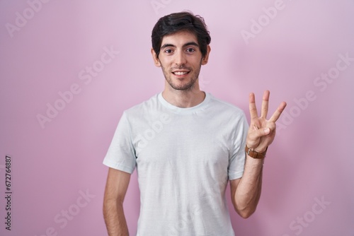 Young hispanic man standing over pink background showing and pointing up with fingers number three while smiling confident and happy.