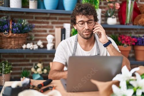 Young hispanic man florist talking on smartphone using laptop at flower shop