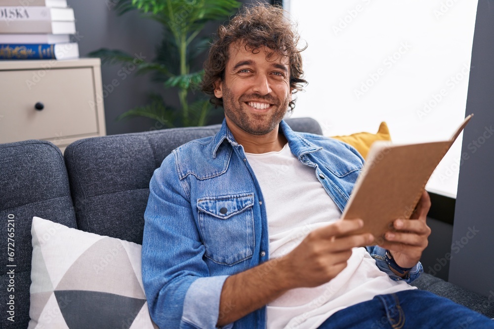 Young hispanic man reading book sitting on sofa at home - obrazy, fototapety, plakaty 